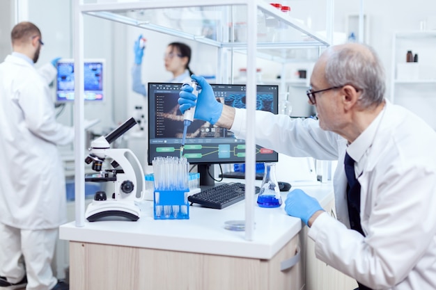 Healthcare specialist in chemistry laboratory doing scientific research with dropper dispenser. Senior professional chemist using pippete with blue solution for microbiology tests.