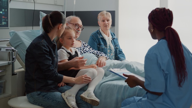 Healthcare specialist doing check up visit with patient and his family in hospital ward. Medical nurse giving assistance and support to elder man with illness to help with recovery.