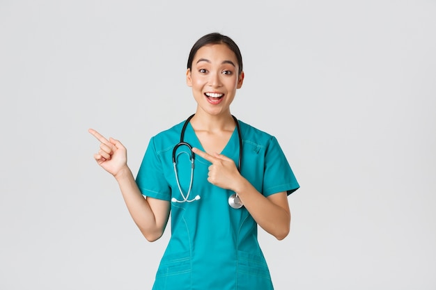 , медицинские работники, концепция пандемии. Взволнованная азиатская медсестра, врач в скрабах и стетоскопе, указывая пальцами в верхнем левом углу. Врач улыбается, удивлен, показывая потрясающее объявление