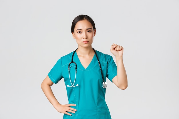 , healthcare workers, pandemic concept. Serious-looking confident serious asian female doctor showing support to fellow collegues during coronavirus, raising fist in unity gesture