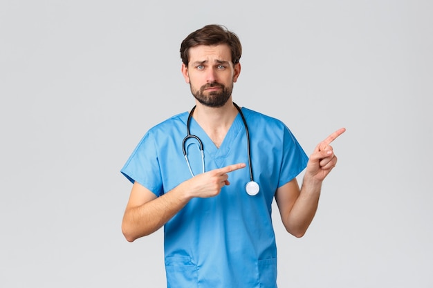 Healthcare workers, pandemic and coronavirus outbreak concept. Unamused and reluctant young doctor in blue scrubs, stethoscope, smirk and frowning skeptical, pointing fingers right