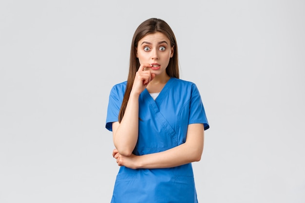 Healthcare workers, prevent virus, insurance and medicine concept. Intrigued female doctor or nurse in blue scrubs having conversation with coworker, biting lip, gossiping, raise eyebrows amused