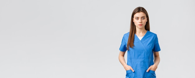 Healthcare workers prevent virus insurance and medicine concept Seriouslooking confident young medical worker nurse or doctor in blue scrubs hold hands in pockets and looking camera