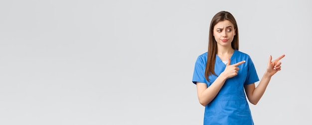 의료 종사자들은 바이러스 보험 및 의학 개념을 예방합니다. 회의적이고 의심스러운 여성 간호사 또는 파란색 수술복을 입은 의사는 웃으면서 판단하는 손가락을 오른쪽으로 응시합니다.