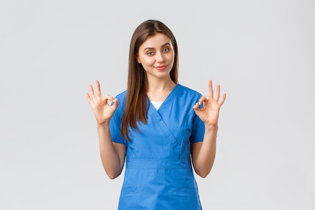 Healthcare workers, prevent virus and medicine concept. Attractive confident female nurse, doctor in blue scrubs, smirk show okay sign, guarantee quality of service in clinic, recommend medicine