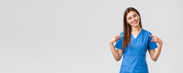 Healthcare workers prevent virus and medicine concept Cheerful smiling pretty doctor female nurse in blue scrubs pointing at herself ready to help patients save lives from coronavirus