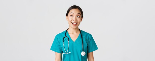 Photo healthcare workers, preventing virus, quarantine campaign concept. enthusiastic smiling asian doctor in scrubs, nurse looking upper left corner with amused face, standing white background.