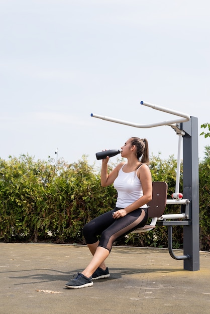 Healthy and active lifestyle. Sports and fitness. Happy woman in white t shirt working out on the sports ground in sunny summer day, drinking water from the thermos bottle