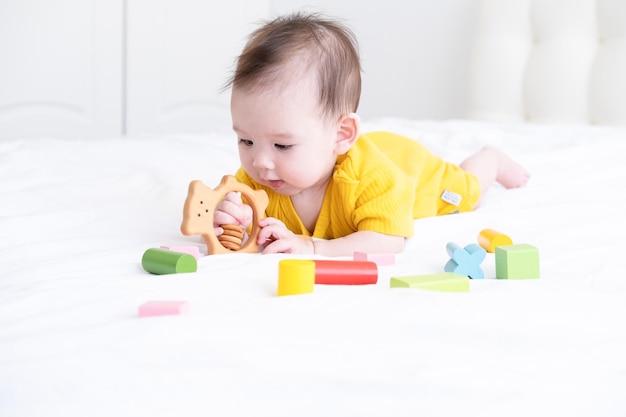白い寝具の上に木製のおもちゃで遊ぶ黄色のボディー スーツで健康なアジアの女の赤ちゃん