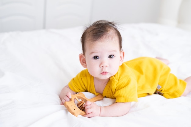 healthy asian baby girl in yellow bodysuit plays with wooden toy teether for teeth on white bedding