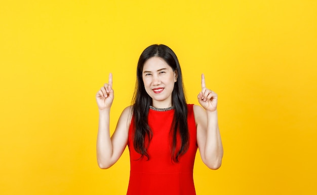 Healthy Asian woman adult wearing beautiful sleeveless dress pointing fingers to high above on bright yellow background.