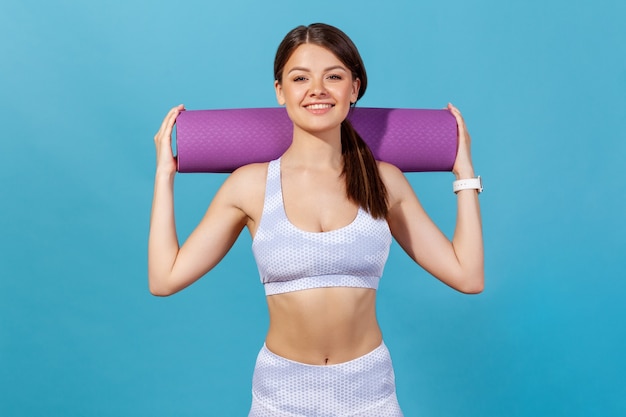 Healthy athletic woman in white sportswear holding rolled mat on shoulders looking at camera