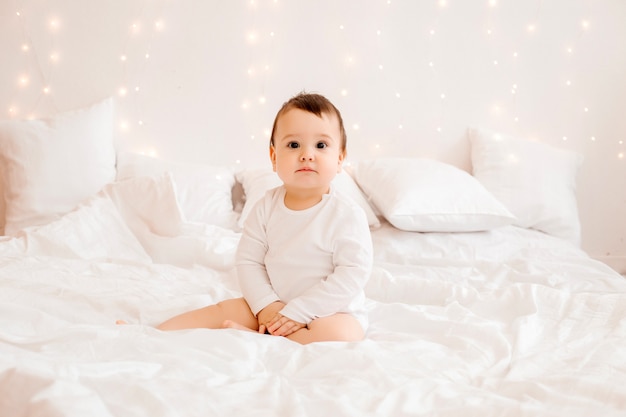 Healthy baby boy 10 months old in white clothes smiling sitting on white bed linen in bed, space for text