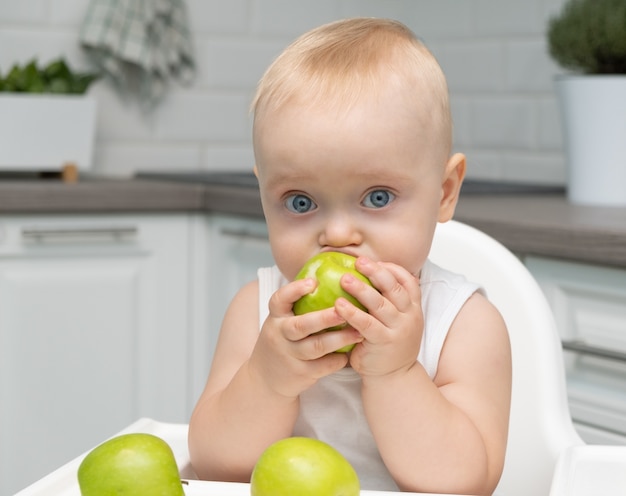Здоровый мальчик с большими голубыми глазами, сидя в детском кресле на кухне, ест зеленые яблоки.