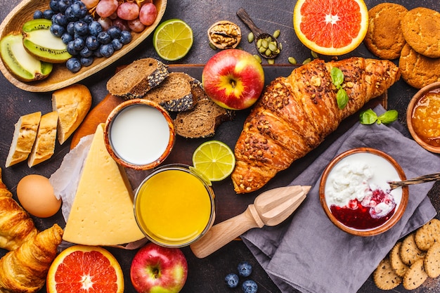 Healthy balanced breakfast on a dark background. Muesli, milk, juice, croissants, cheese, biscuits.