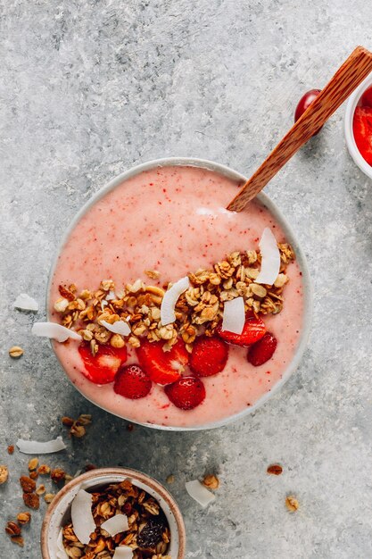 Healthy banana and strawberrie smoothie in a bowl with pieces of strawberries granola and coconut chips on light gray background Top view