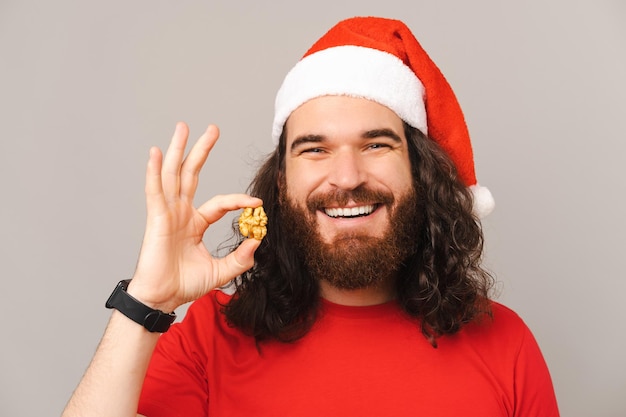 Un uomo barbuto sano che indossa un cappello di natale tiene in mano una noce vicino al viso