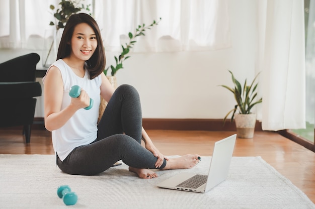 Healthy beautiful Asian woman sitting on the floor holding dumbbell using laptop at home in living room ready to online workout