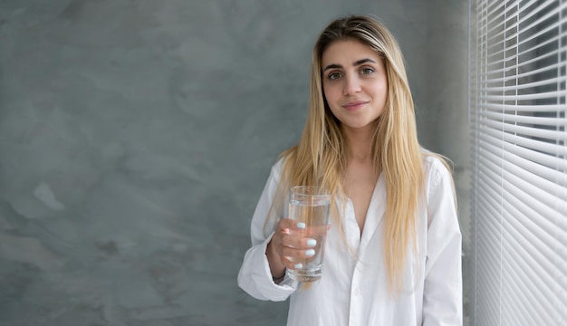 Bella giovane donna in buona salute si trova a casa vicino alla finestra con in mano un bicchiere d'acqua