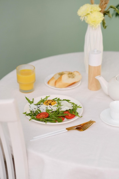 Healthy breakfast of fried egg with tomatoes and green on white plate orange juice bread teapot and