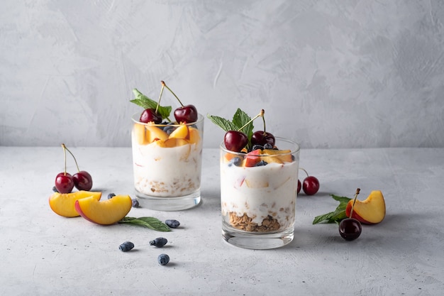 Healthy breakfast in a glass with fresh fruits: pomegranate, cherry, nectarines, honeysuckle, yogurt and granola on a grey background. Shallow depth of field with selective focus
