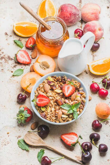 Healthy breakfast ingredients Oat granola in bowl with nuts strawberry and mint milk in pitcher honey in glass jar fresh fruits berries on light concrete background