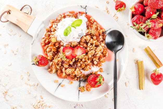 Photo healthy breakfast. oatmeal granola crumble with rhubarb, fresh strawberries and blackberries, seeds and ice cream on marble white plate, with mint, on white stone concrete table,  top view