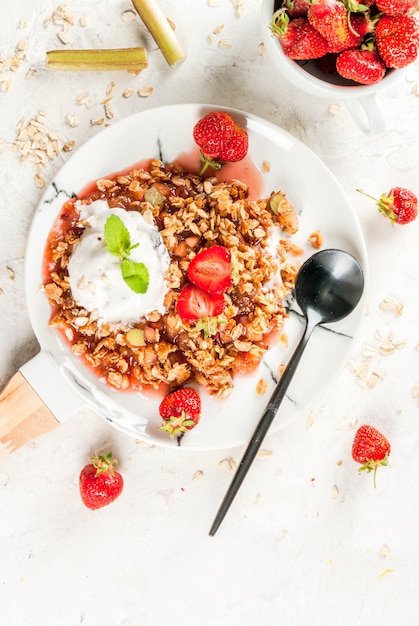 Healthy breakfast. Oatmeal granola crumble with rhubarb, fresh strawberries and blackberries, seeds and ice cream on marble white plate, with mint, on white stone concrete table,  top view