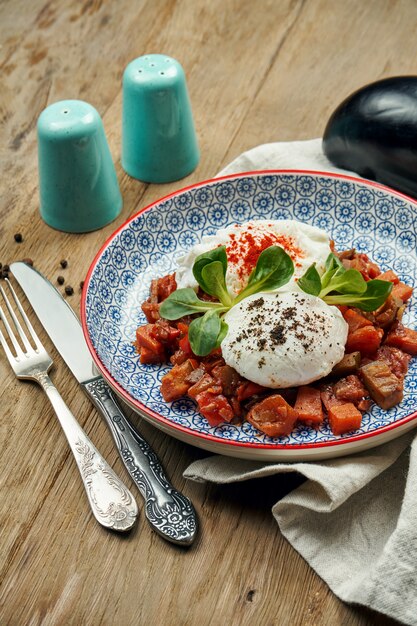 Healthy breakfast - poached egg with stewed vegetables and yogurt sauce in a blue bowl on a wooden surface. Cilbir