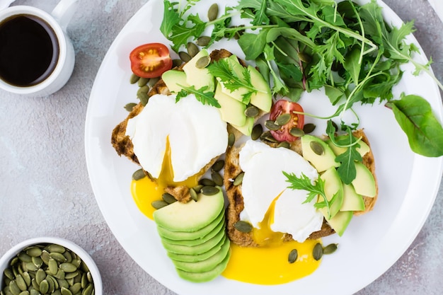 Colazione salutare. uova in camicia su pane tostato con pezzi di avocado, rucola, mizuna e foglie di bietola e pomodori su un piatto su un tavolo servito. dieta flexetaria. vista dall'alto. avvicinamento