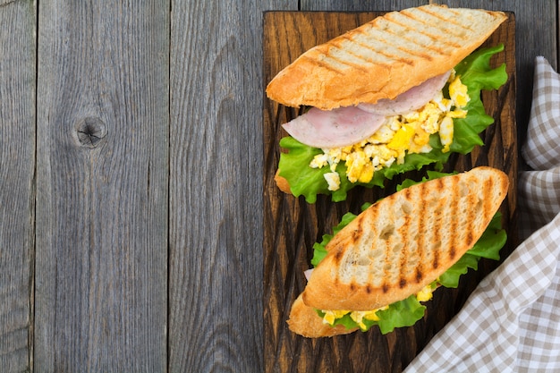 Healthy breakfast. Sandwich stuffed with scrambled eggs, ham and lettuce leaves on old wooden background. Top view. Selective focus.