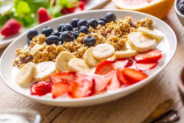 Здоровый завтрак подается с тарелкой клубники черники мюсли йогурта и банана. Утренний стол мюсли, миндаль, ягоды, сок цитрусовых и зелень.