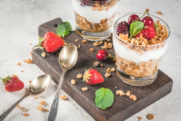 Foto colazione salutare. frutti e bacche estive. yogurt greco fatto in casa con muesli, more, fragole, ciliegie e menta. sul tavolo di pietra bianca di cemento, in bicchieri.