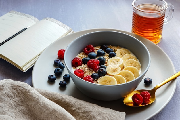 Sana colazione con farina d'avena in una ciotola, fette di banana, lampone, mirtillo, una tazza di tè e un taccuino con una penna sul piano del tavolo grigio chiaro