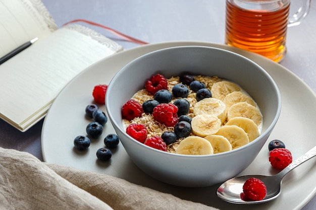 Sana colazione con farina d'avena in una ciotola, fette di banana, lampone, mirtillo, una tazza di tè e un taccuino con una penna sul piano del tavolo grigio chiaro