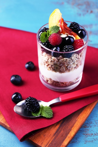 Здоровый йогурт для завтрака со свежими фруктовыми ягодами и мюсли подается в стекле на цветном деревянном фоне
