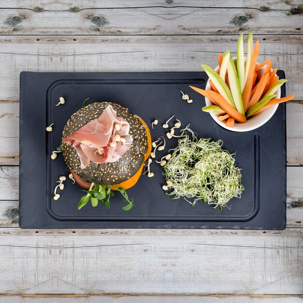 Healthy burger with hamon, tomatoes, micro greens and black wholegrain buns, vegetable sticks on black slate board over wooden table. Clean eating, dieting, detox food concept.