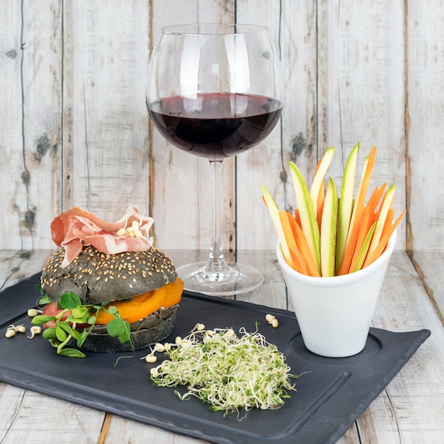 Healthy burger with hamon, tomatoes, micro greens and black wholegrain buns, vegetable sticks and red wine on black slate board