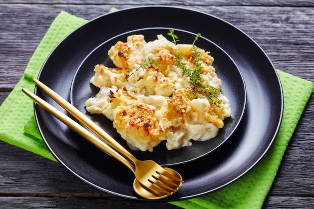 Healthy casserole cauliflower cheese served on a black plate with fresh thyme on top on wooden table with green napkin and golden cutlery, close-up