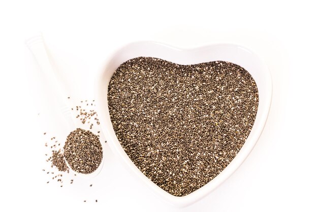 Здоровые семена чиа в миске в форме сердца на белом фоне.