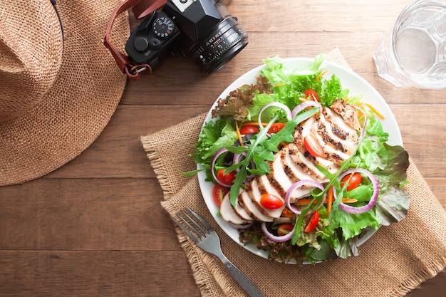 Healthy chicken salad with mixed green and tomatoes on wooden table with hat and camera. Healthy