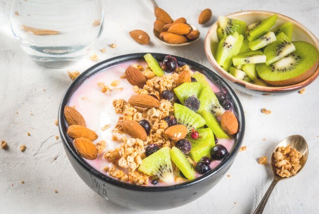 Photo healthy diet breakfast: smoothies bowl, with yogurt, fresh blueberries black currant, kiwi, granola oatmeal, almond nuts. on a white stone table, with a glass of water and ingredients.