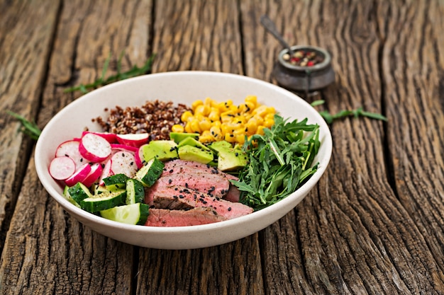 Healthy dinner. Buddha bowl lunch with grilled beef steak and quinoa, corn, avocado, cucumber and arugula on wooden table. Meat salad.