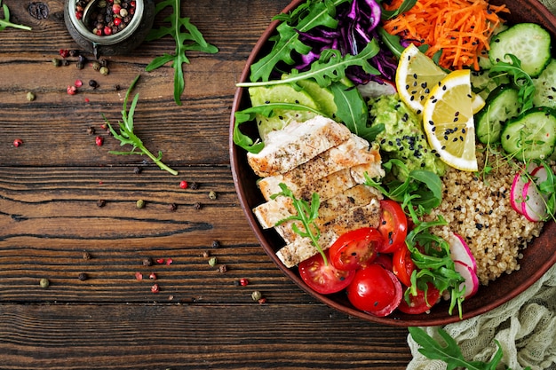 Healthy dinner. Buddha bowl lunch with grilled chicken and quinoa, tomato, guacamole, carrot, red cabbage, cucumber and arugula on wooden table. Flat lay. Top view