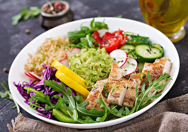 Healthy dinner. Buddha bowl lunch with grilled chicken and quinoa, tomato, guacamole, red cabbage, cucumber and arugula on gray table.