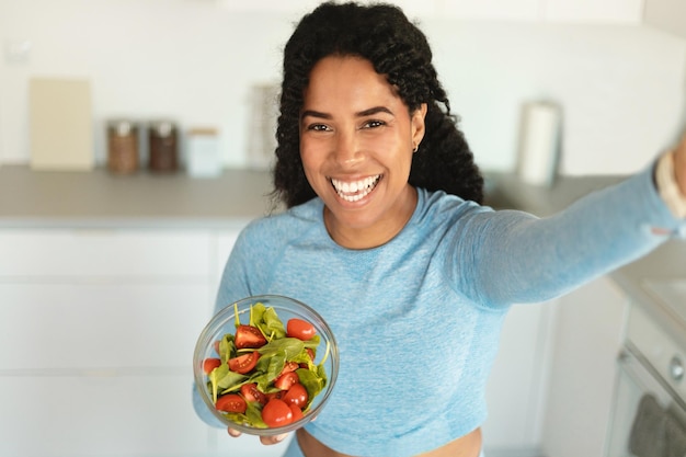 Healthy eating concept Happy sporty black lady holding bowl with fresh vegetable salad and taking selfie in kitchen