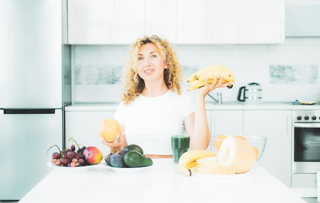 Концепция здорового питания улыбающаяся молодая женщина держит банановые и апельсиновые фрукты на кухне, ест и лечится