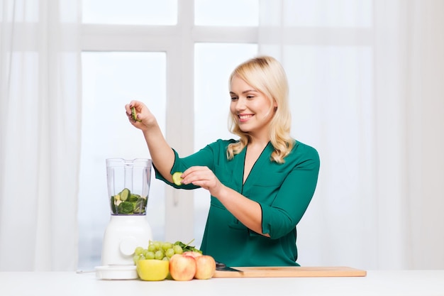 healthy eating, cooking, vegetarian food, diet and people concept - smiling young woman putting fruits and vegetables into blender at home kitchen