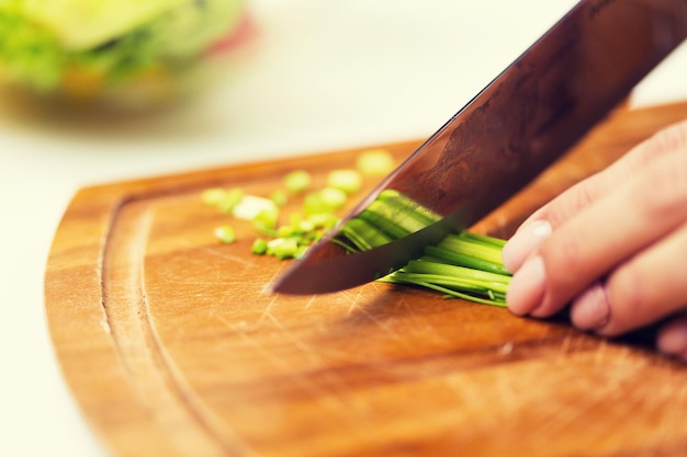 healthy eating, cooking, vegetarian food, dieting and people concept - close up of woman chopping green onion with knife on wooden cutting board