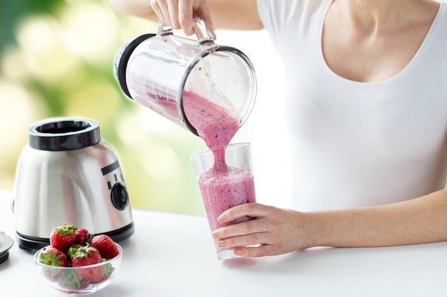 healthy eating, cooking, vegetarian food, dieting and people concept - close up of woman with blender and strawberries pouring milk shake to glass over green natural background